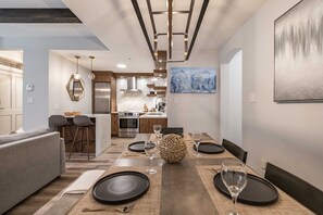 Une cuisine moderne équipée d’appareils électroménagers en acier inoxydable, un coin repas avec une table en bois pour quatre personnes et un salon avec un canapé et des fauteuils.
