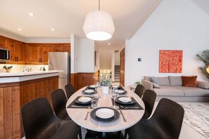 On aperçoit un coin repas avec une table ronde dressée pour six personnes, entourée de chaises. Au fond, on distingue une cuisine avec des meubles en bois et un réfrigérateur.