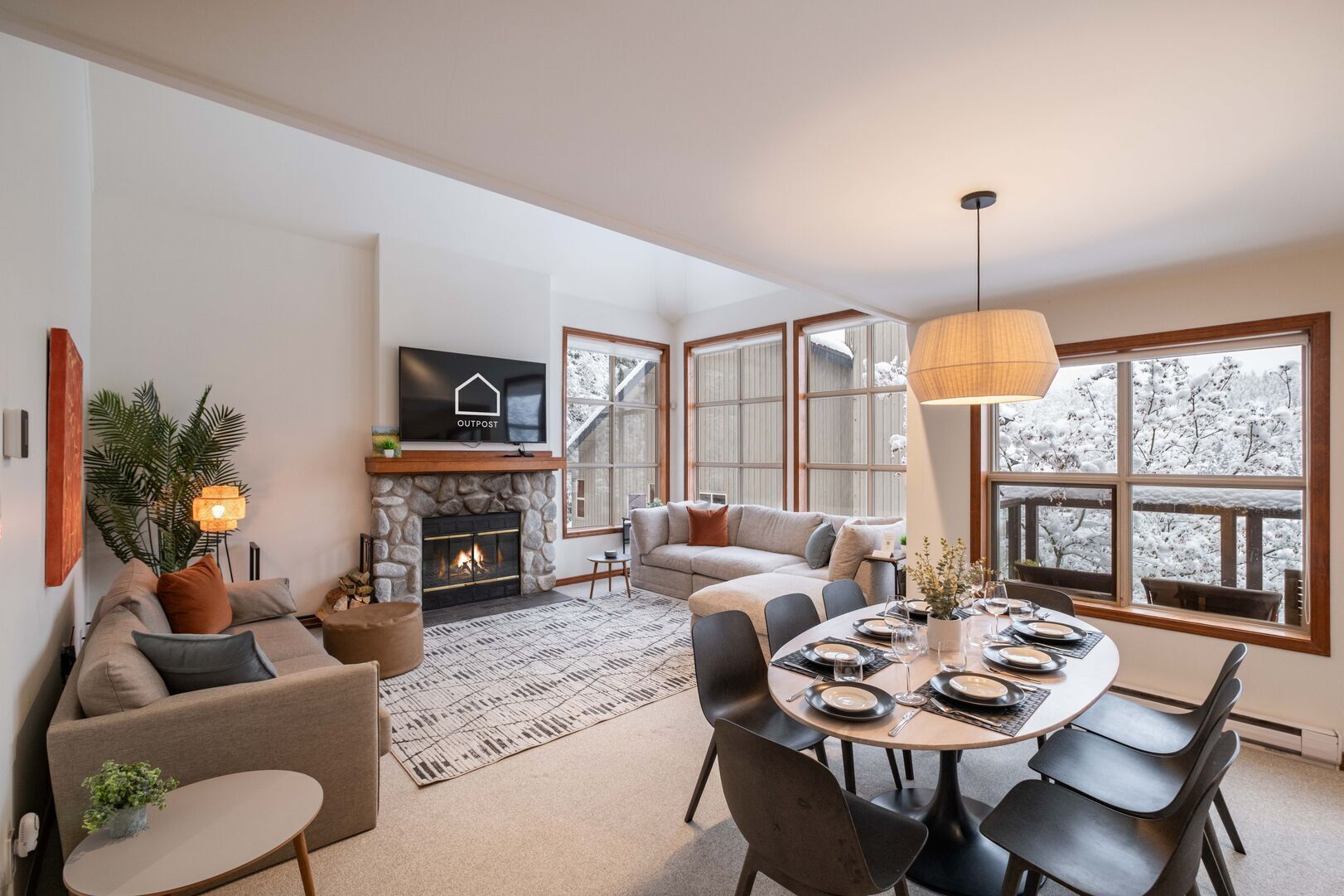 Ein modernes Wohnzimmer mit Kamin, Fernseher, Essbereich mit Tisch für vier Personen und großen Fenstern mit Blick auf schneebedeckte Bäume.