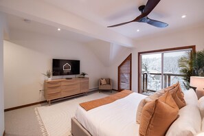 Ein Schlafzimmer mit einem Bett, einem Fernseher auf einer Holzkommode, einem Deckenventilator und einem großen Fenster mit Blick auf schneebedeckte Bäume.