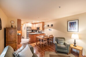 A cozy living room with a sofa, a dining table with chairs, a kitchen area with wooden cabinets, and a small side table with a lamp. - Close to Blackcomb | Shared hot tub | Free parking (Whistler)