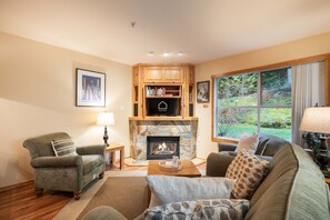 A cozy living room with a fireplace, wooden bookshelves, and a large window offering a view of the outdoors. - Close to Blackcomb | Shared hot tub | Free parking (Whistler)