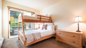 Una cama litera con estructura de madera, una mesita de noche con lámpara y vistas a un jardín a través de una puerta de cristal.