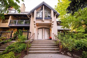 A multi-story building with a prominent entrance, a staircase leading to the main door, and a well-maintained garden area. - Benchlands Townhome | Golf, Ski & Free Parking (Whistler)