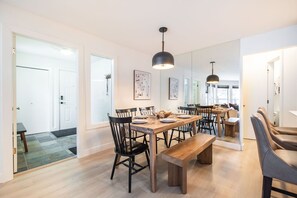 Ein geräumiger Essbereich mit einem langen Holztisch, schwarzen Stühlen und einer Bank. Über dem Tisch hängen Pendelleuchten, und der Raum ist modern und minimalistisch eingerichtet, mit weißen Wänden und Holzböden.