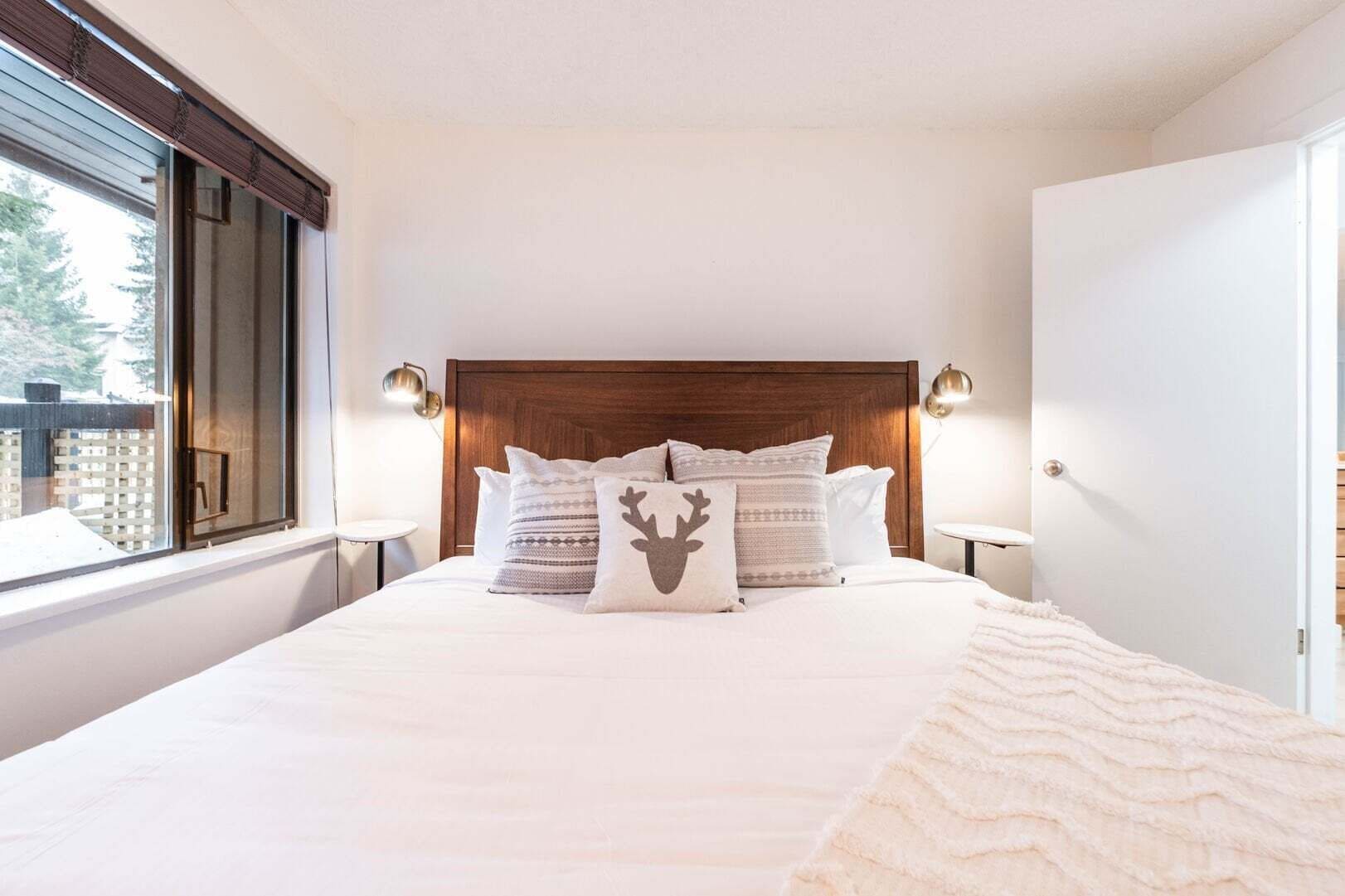 A bedroom with a large bed, a wooden headboard, two bedside lamps, and a window with a view of trees.