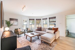 Ein modernes Wohnzimmer mit einer Sofagarnitur, einem Couchtisch und einer Lampe. Große Fenster mit Jalousien, eine Pflanze und eine Tür zu einem weiteren Raum sind vorhanden.