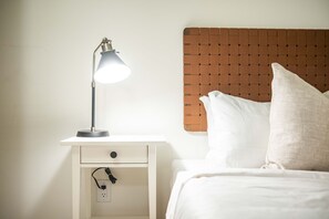 Ein ordentlich bezogenes Bett mit weißer Bettwäsche und Kissen, eine Nachttischlampe auf einem weißen Nachttisch und eine Steckdose.