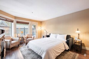 Ein Schlafzimmer mit einem Bett, zwei Stühlen, einem Schreibtisch, einer Lampe und Blick durch große Fenster.