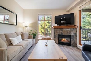 Ein Wohnzimmer mit einer Couch, einem Couchtisch, einem Kamin, einem Fernseher und einem Balkon mit Blick auf Bäume.