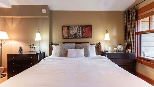 Quarto de hotel com uma cama grande, duas mesas de cabeceira com luminárias, um quadro na parede e uma janela com cortinas.