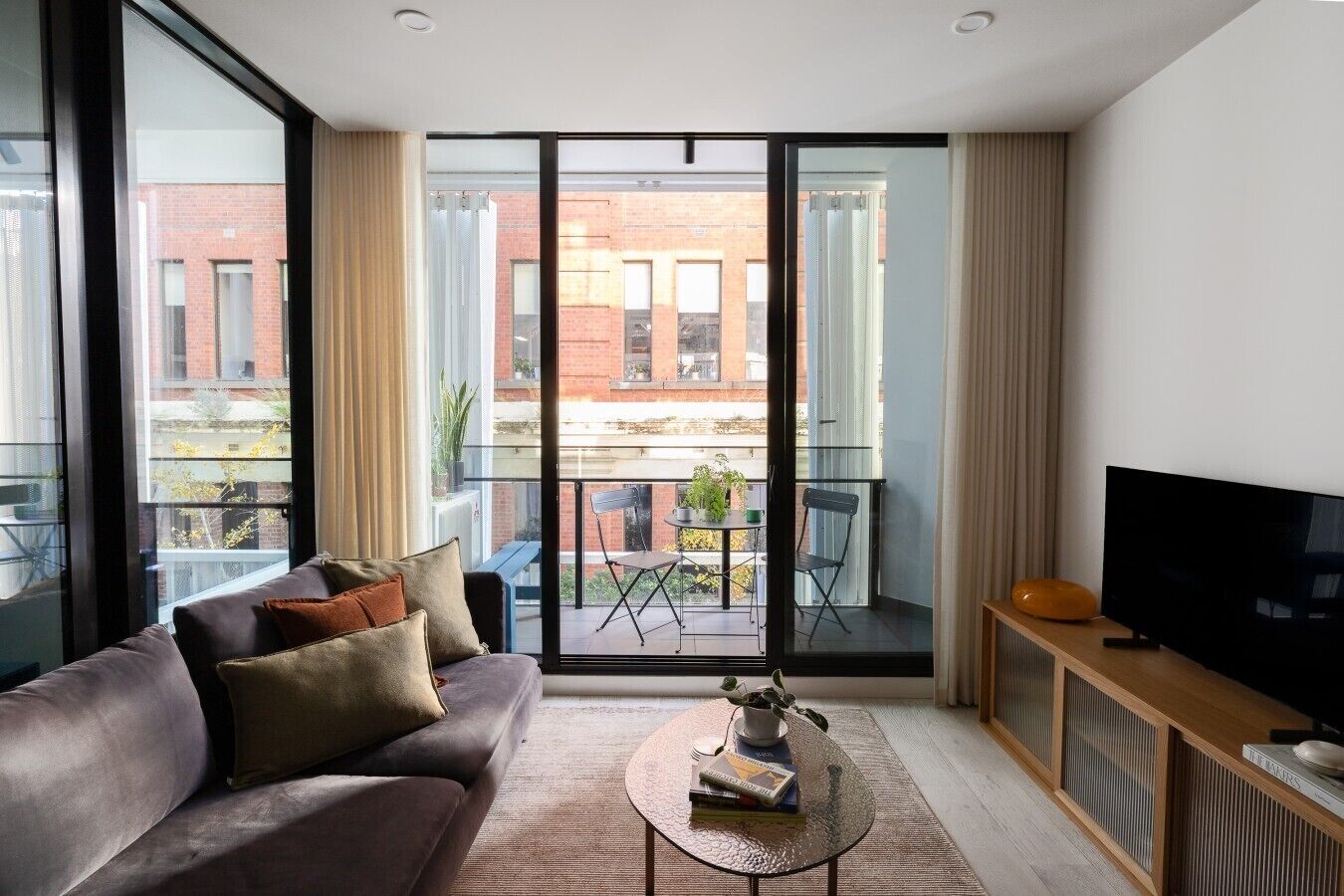 Een moderne woonkamer met een bank, een salontafel, een televisie en een balkon met uitzicht op een bakstenen gebouw.