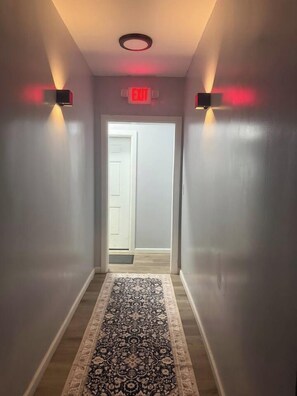 A hallway with a patterned rug, a door at the end, and wall-mounted lights. - Renovated 3BR | Bay Windows | EWR 15 Min (East Orange)
