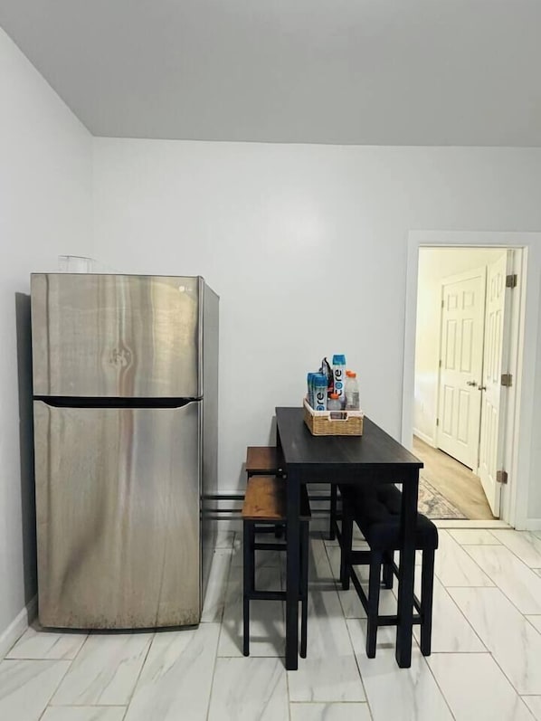 A kitchen with a stainless steel refrigerator, a wooden dining table with chairs, and a basket of bottled water. - Renovated 3BR | Bay Windows | EWR 15 Min (East Orange)