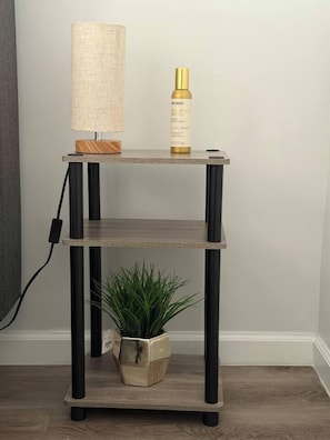 A wooden console table with a lamp and a bottle on top, a potted plant below, and a power cord on the left. - Renovated 3BR | Bay Windows | EWR 15 Min (East Orange)