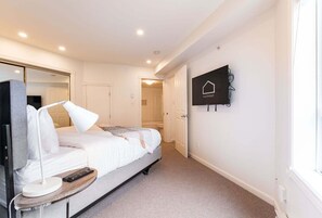 A hotel room with a bed, bedside table, lamp, remote control, and a mounted television. - NEW Townhome | Private hot tub | Complex pool (Whistler)