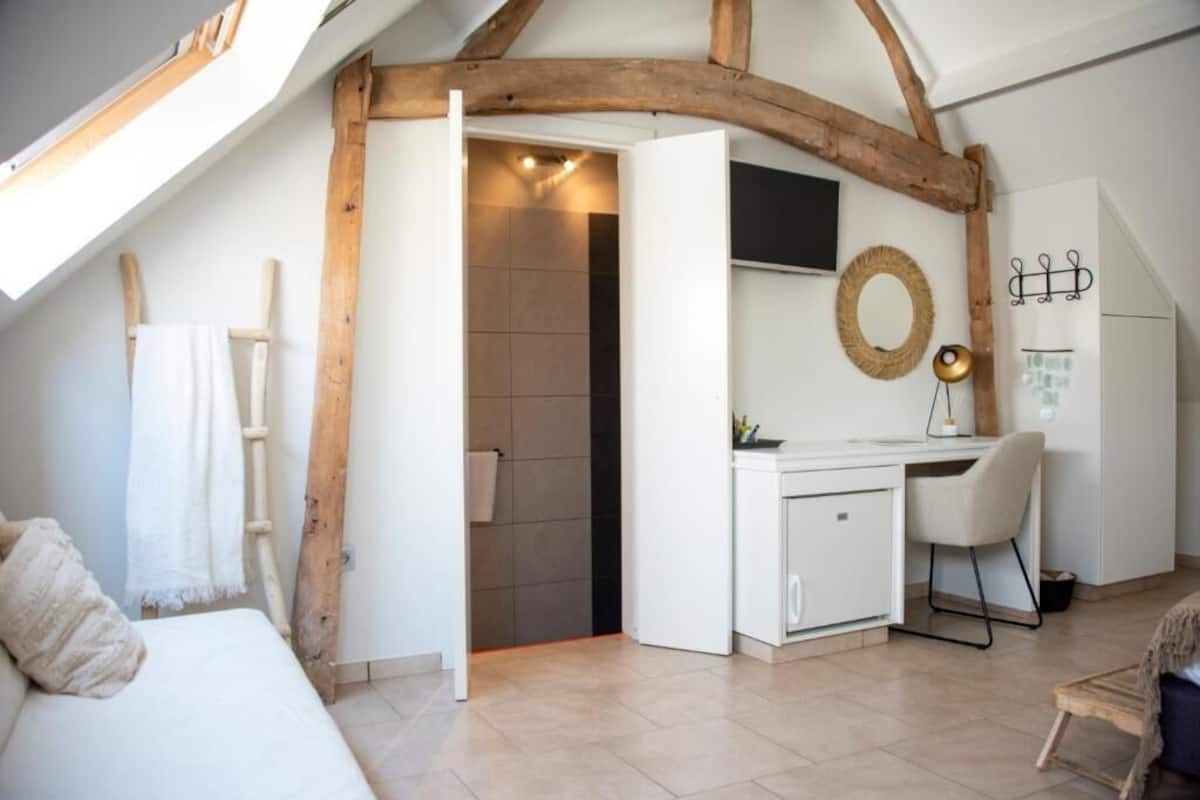 A room with a sloped ceiling, wooden beams, a white desk, a chair, a bed, and a small bathroom.
