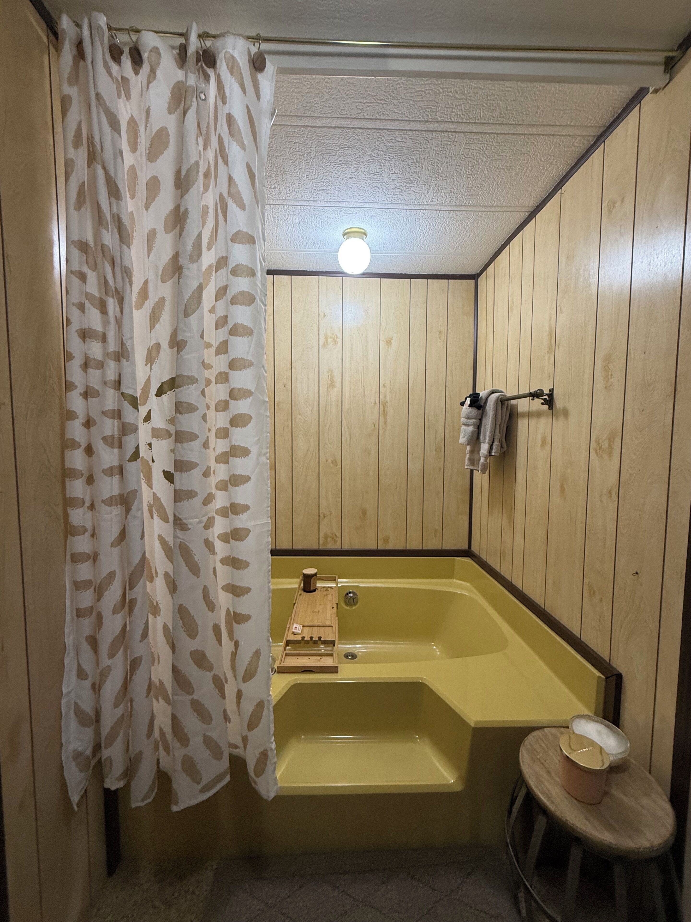 A small bathroom with a wooden panel wall, a bathtub, a shower curtain, a towel rack, and a small stool.