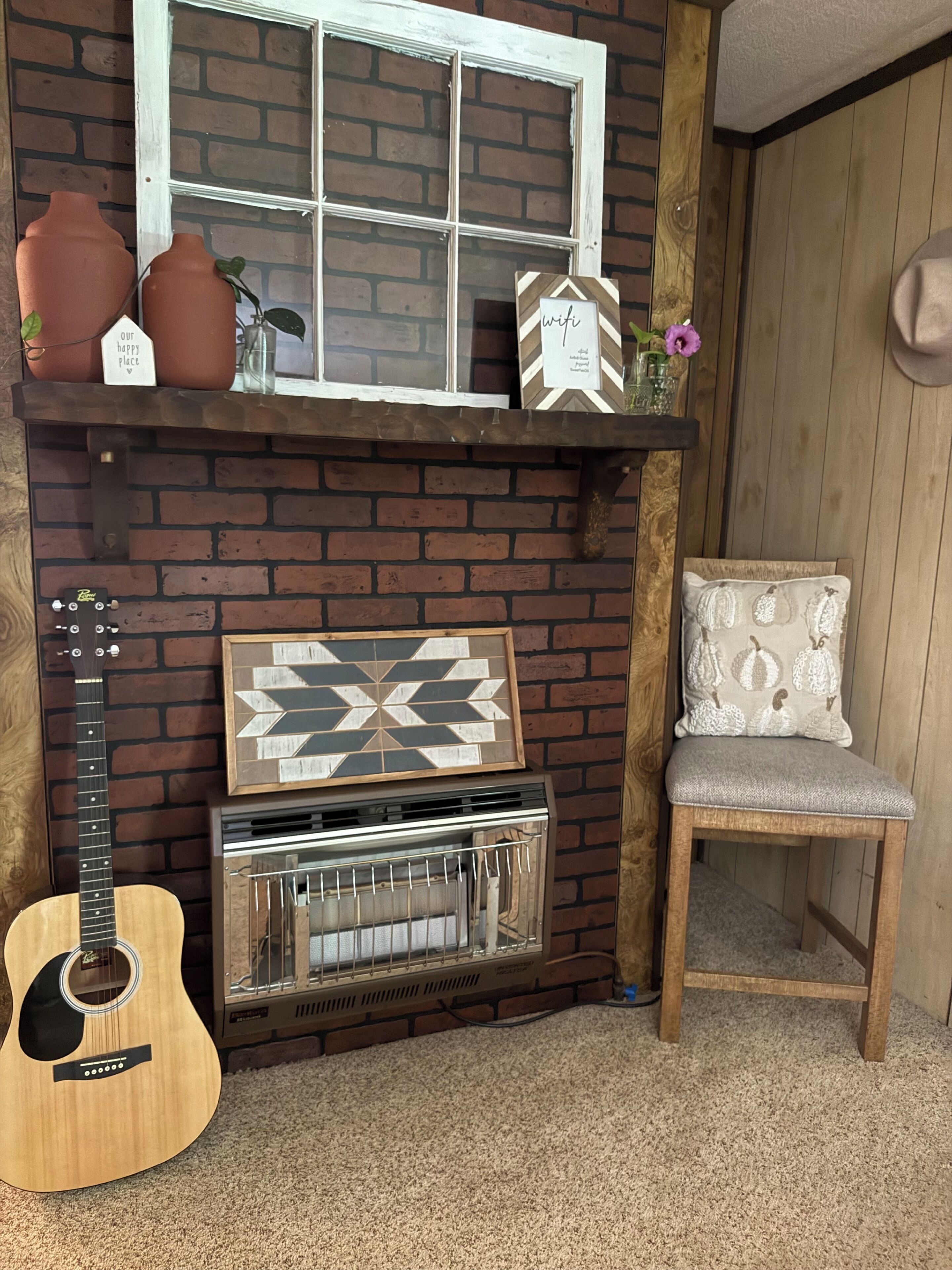 Une cheminée en briques surmontée d’une étagère où sont exposés divers objets décoratifs, une guitare et un fauteuil avec un coussin.