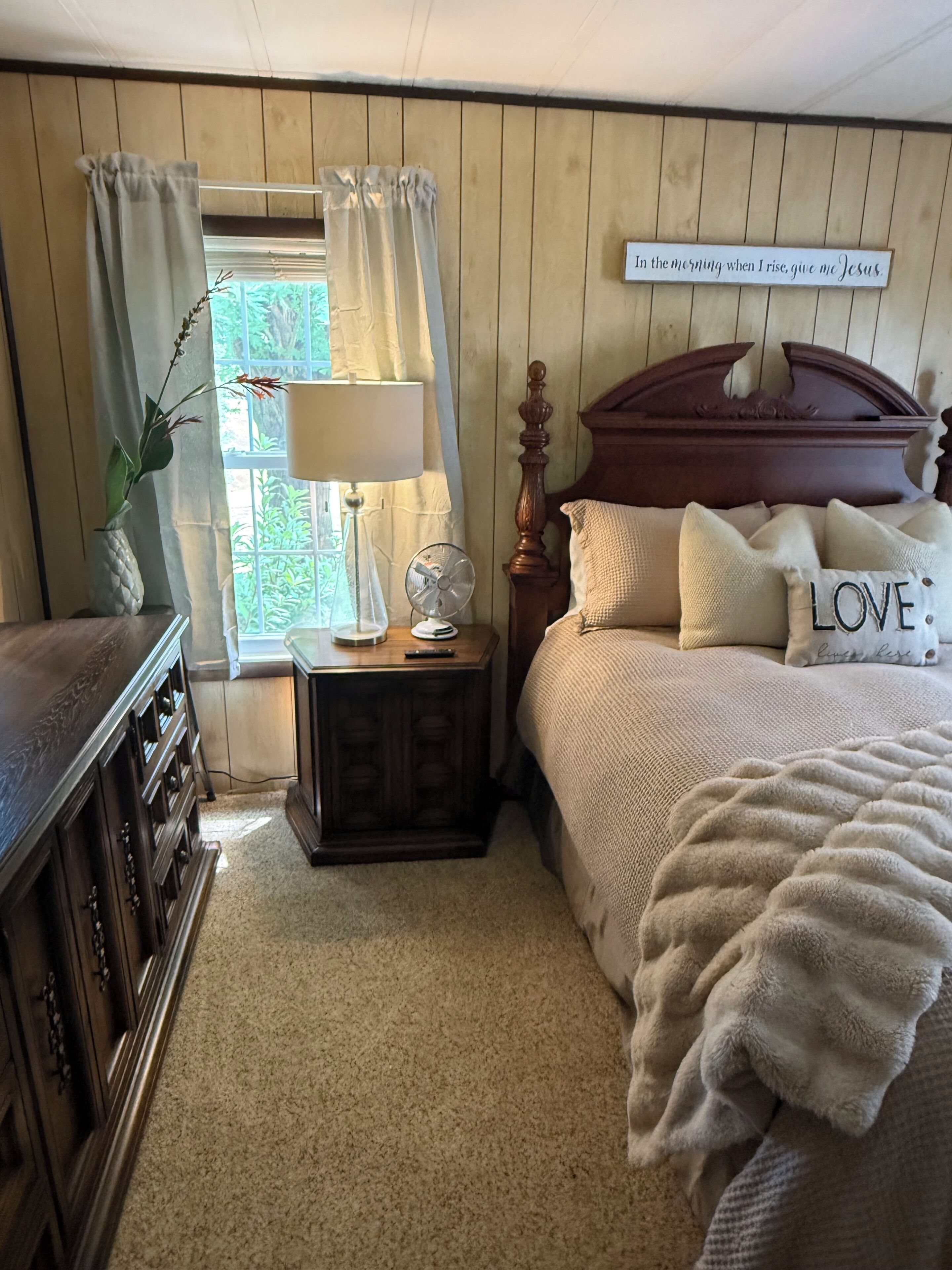 Une chambre à coucher avec une tête de lit en bois, une table de chevet avec une lampe, un ventilateur et une fenêtre avec des rideaux.