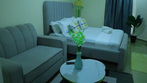Una camera d'albergo con un divano grigio, un letto con biancheria bianca, un vaso con fiori, una lampada e una pianta in vaso.