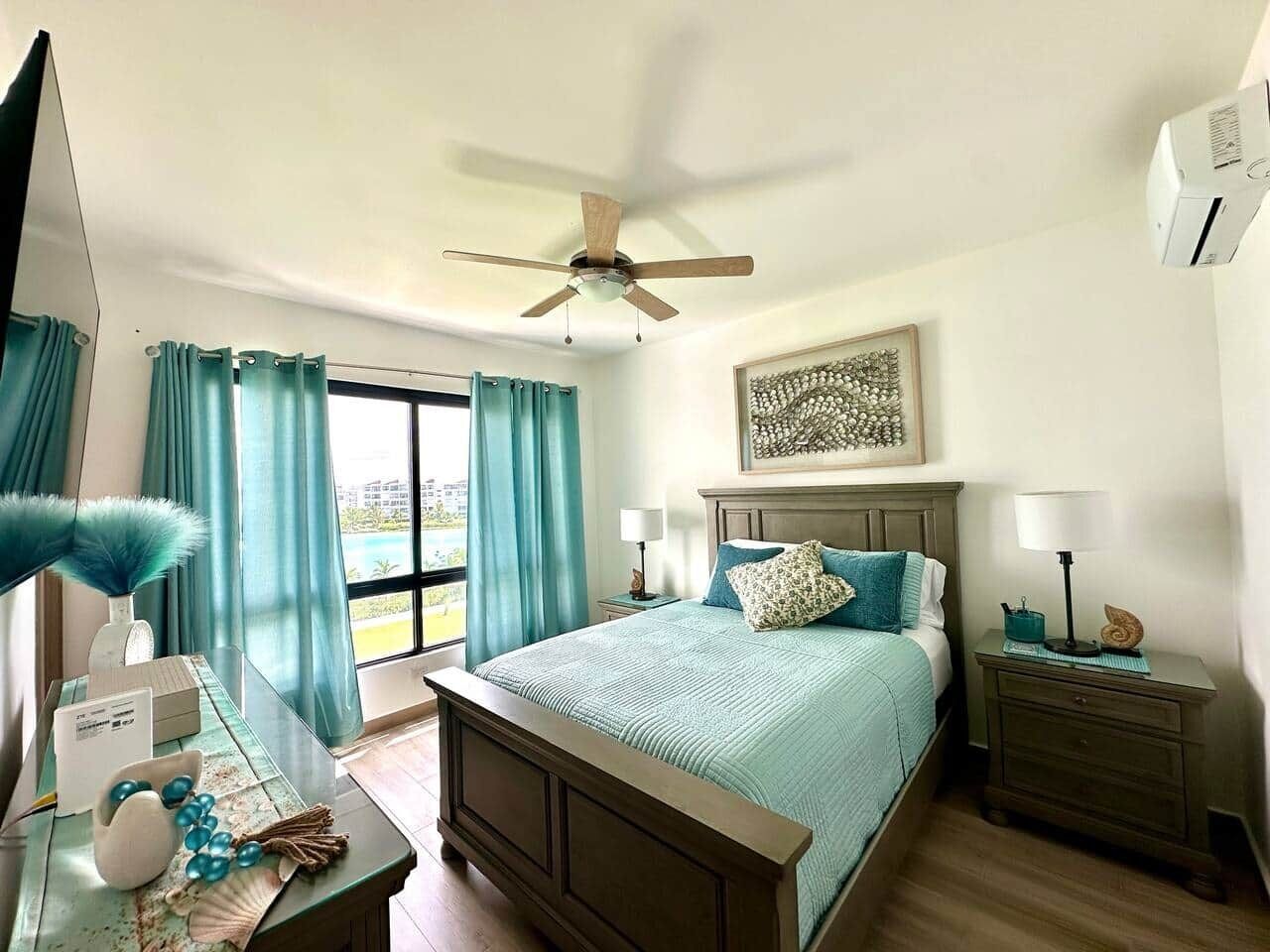 Habitación de hotel con cama, escritorio con lámpara, ventilador de techo y ventana con cortinas.