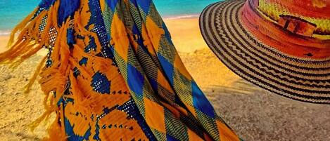 A hammock hanging under a thatched roof, a colorful woven blanket, and a straw hat on a beach with clear blue water.