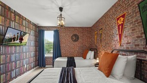 Ett hotellrum med en tegelvägg, en bokhylla, två sängar, en tv och ett fönster med gardiner.