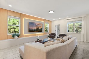 A modern living room with a large sofa, a coffee table, and a TV mounted on the wall. There are windows with curtains, a door leading to a bathroom, and a potted plant on the windowsill. - Resort Amenities Included! 7 BD/ 6 BA/ Storey Lake 4585 TB (Kissimmee)