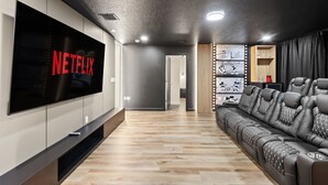 A modern living room with a large flat-screen TV, a leather sofa, and a wall-mounted shelf with decorative items. - Resort Amenities Included! 7 BD/ 6 BA/ Storey Lake 4585 TB (Kissimmee)