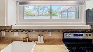 A kitchen with white cabinets, a window with a view of palm trees, a tiled backsplash, and a stove. - Beach House Oasis - Steps from Ocean Access (Venice)