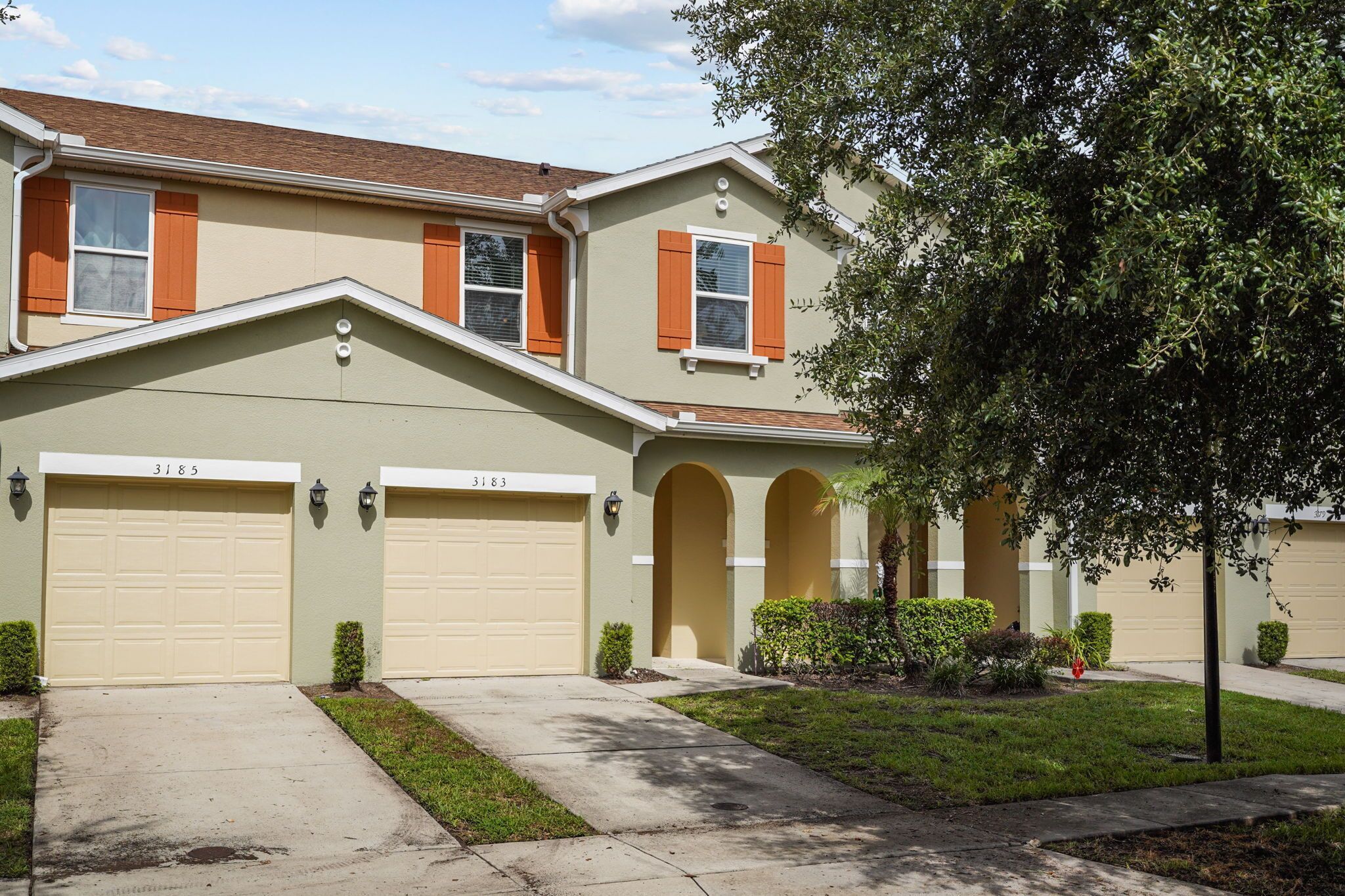 A two-story residential building with two garage doors, a small front yard, and a tree.
