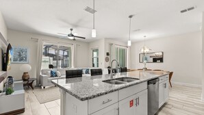 A modern kitchen with a large island, a dining area with a table and chairs, and a living room with a sofa and a ceiling fan. - 4 BD/ 3 BA/ Storey Lake (3071 GS) (Kissimmee)
