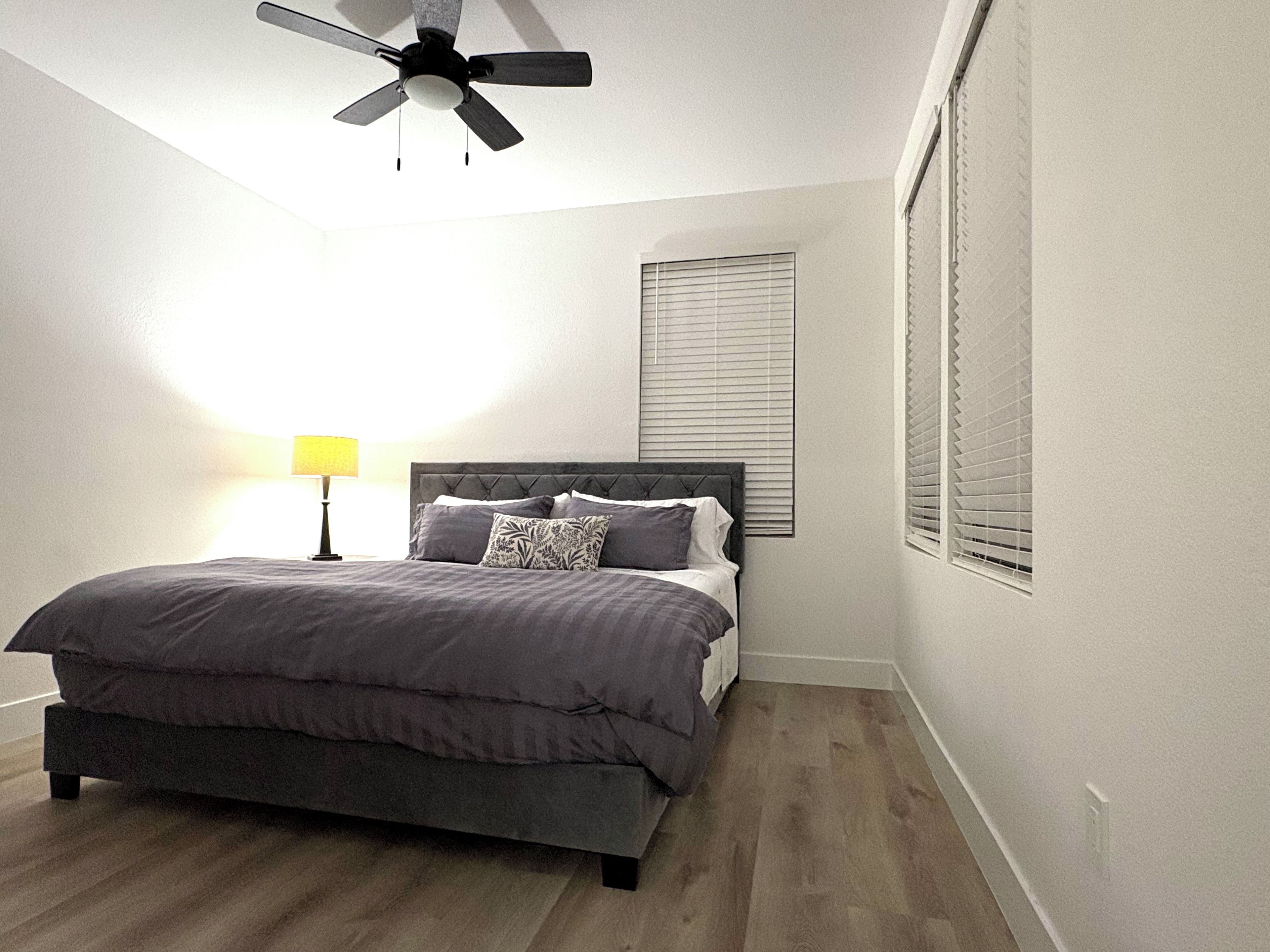 Ein Schlafzimmer mit einem Bett, einem Deckenventilator, einer Nachttischlampe und einem Fenster mit Jalousien.