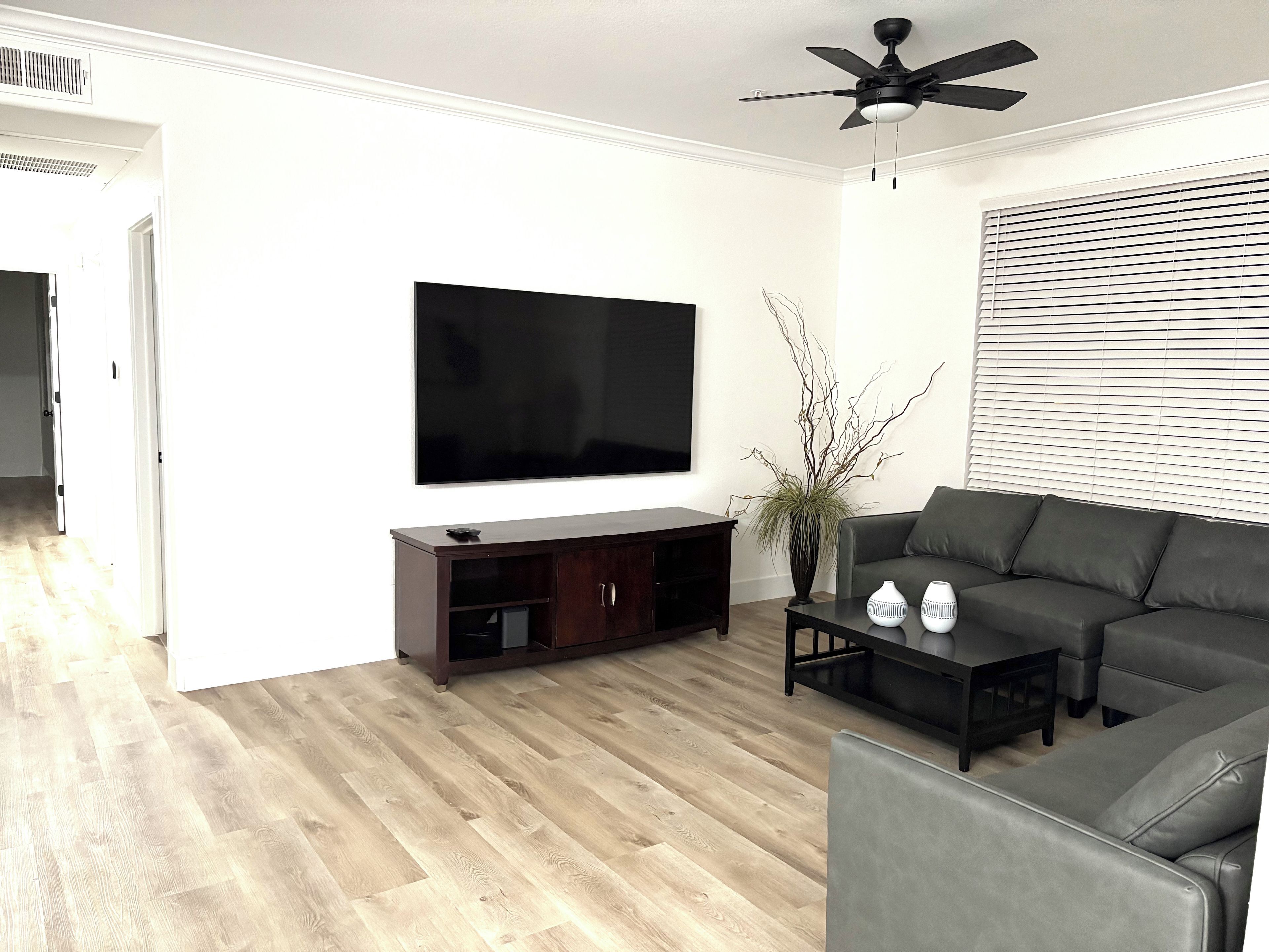 A living room with a flat-screen TV, dark wooden entertainment unit, grey sofa, and a ceiling fan.