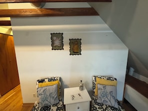 Una stanza con un muro bianco, due poltrone a fantasia, un piccolo tavolo bianco con una bottiglia e un soffitto con travi in legno.