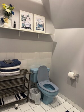 A bathroom with a toilet, a towel rack with folded towels, a trash bin, and two framed artworks on the wall. - Cozy 1 bed,  furnished apartment (Chicago)