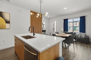 A modern kitchen with a central island, dining area with chairs, and a view of a building through large windows. - 3 BD/ 3 BA/ The Hub at Westside Reserve (3263 SW) (Kissimmee)