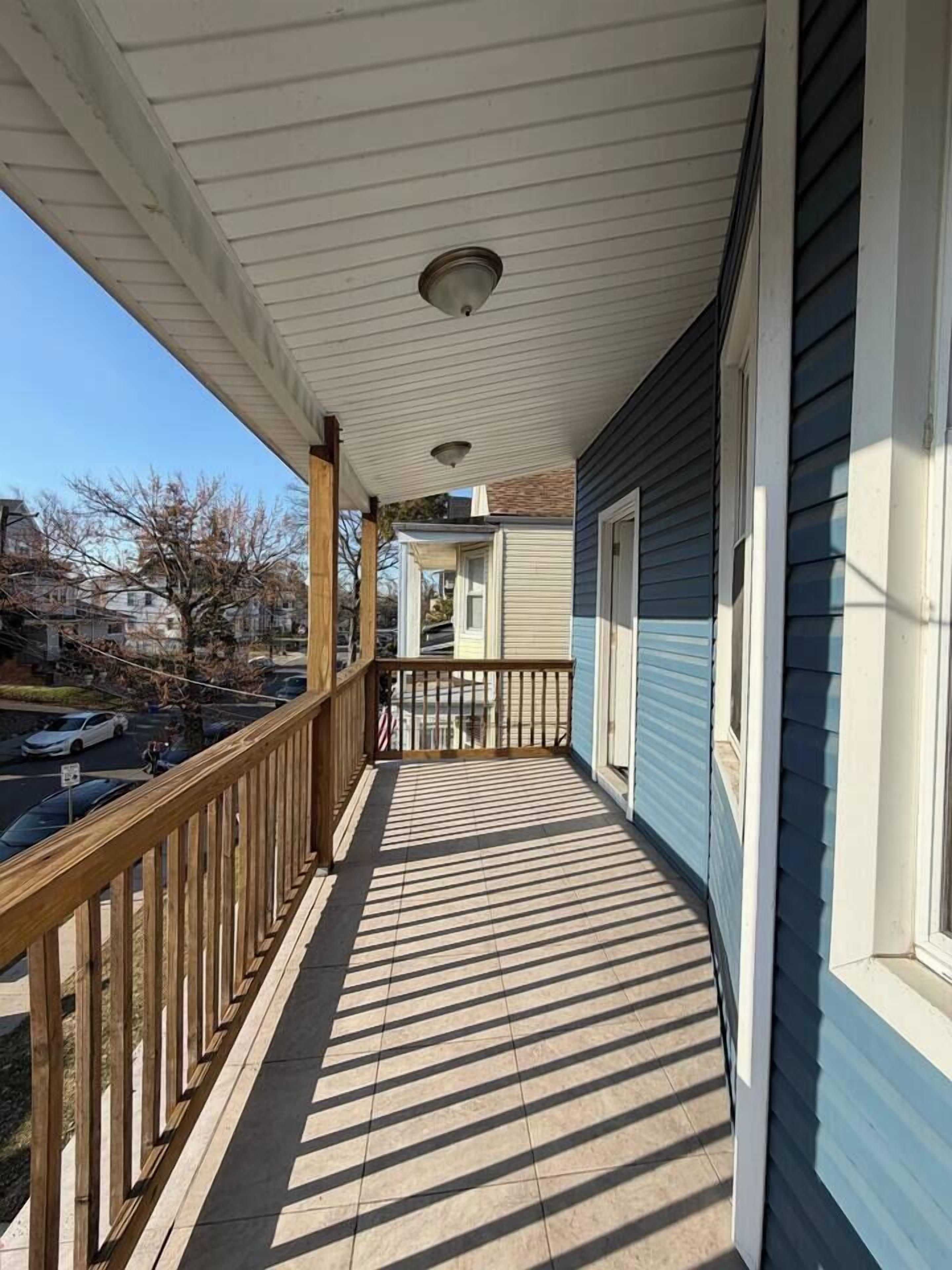 A covered porch with a wooden railing, a tiled floor, and a view of a street and houses in the background.