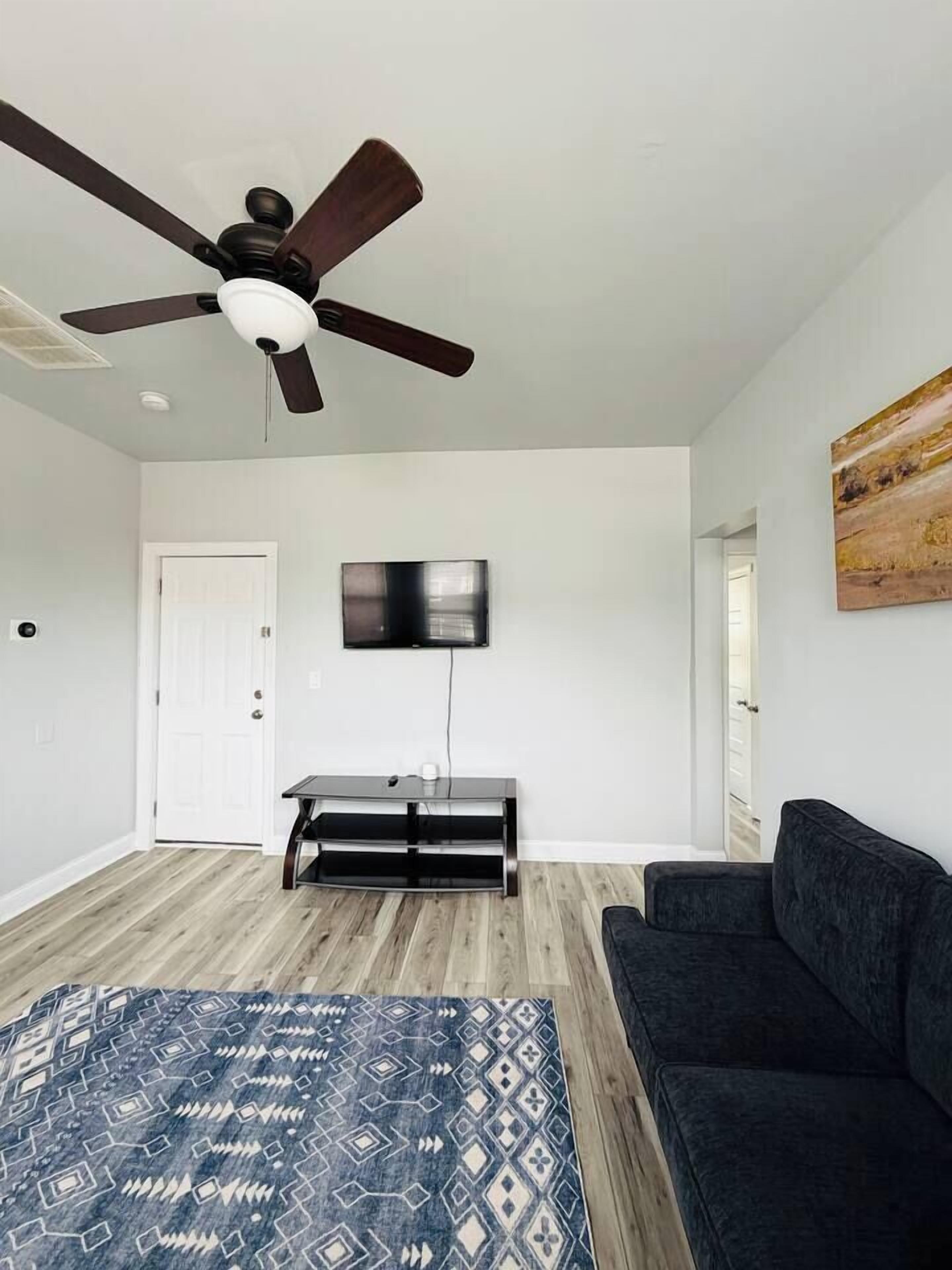 A living room with a ceiling fan, a flat-screen TV mounted on the wall, a dark sofa, a patterned rug, and a wooden floor.