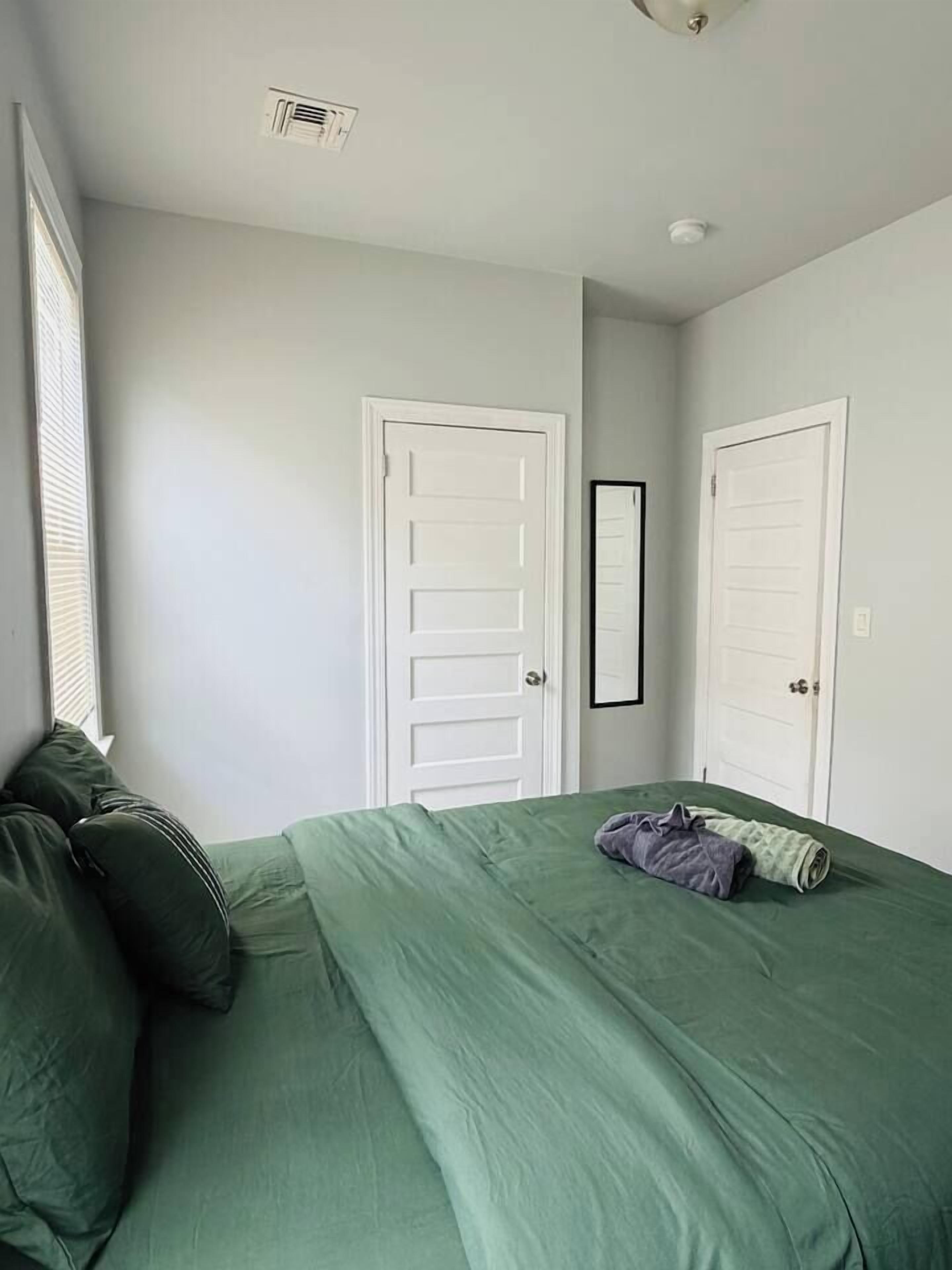 A bedroom with a green bedspread, two white doors, a window with blinds, and a ceiling vent.