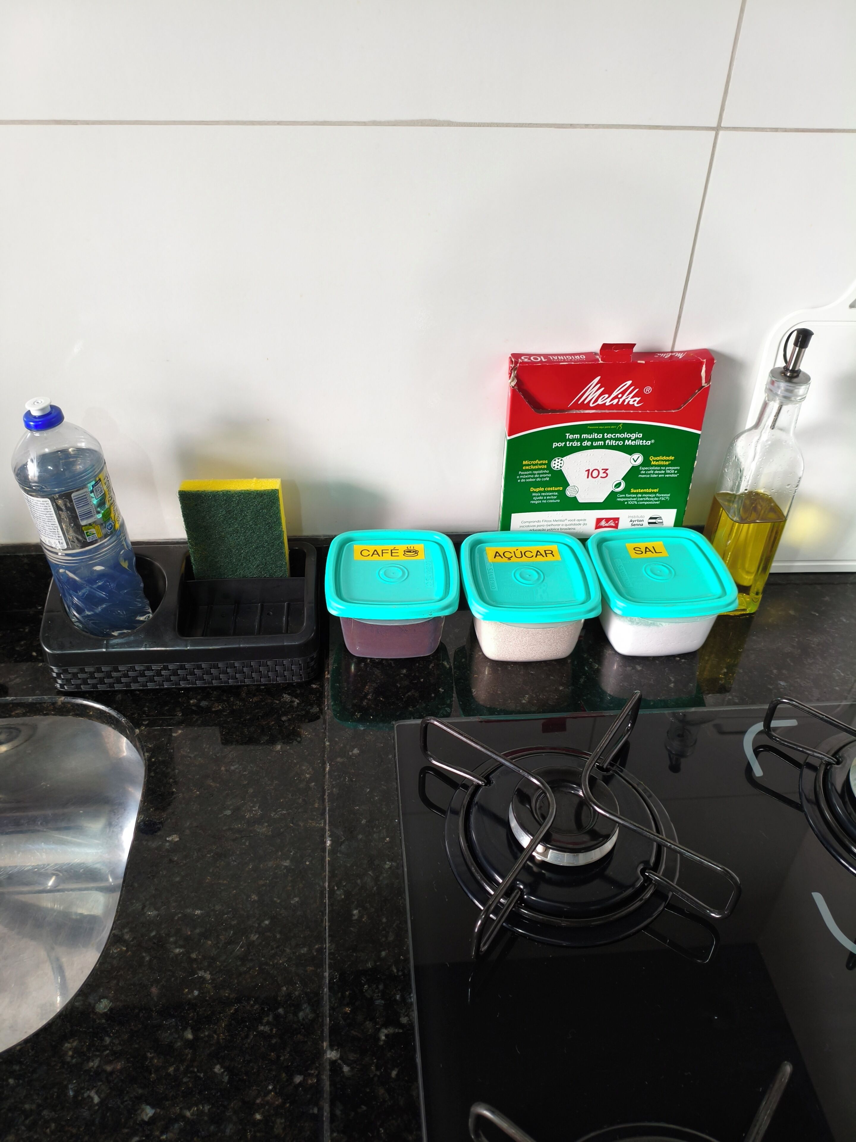 Un comptoir de cuisine avec une bouteille d’eau, une éponge, une boîte de savon Melita et des récipients étiquetés « CAFÉ », « SUCRE » et « SEL ».