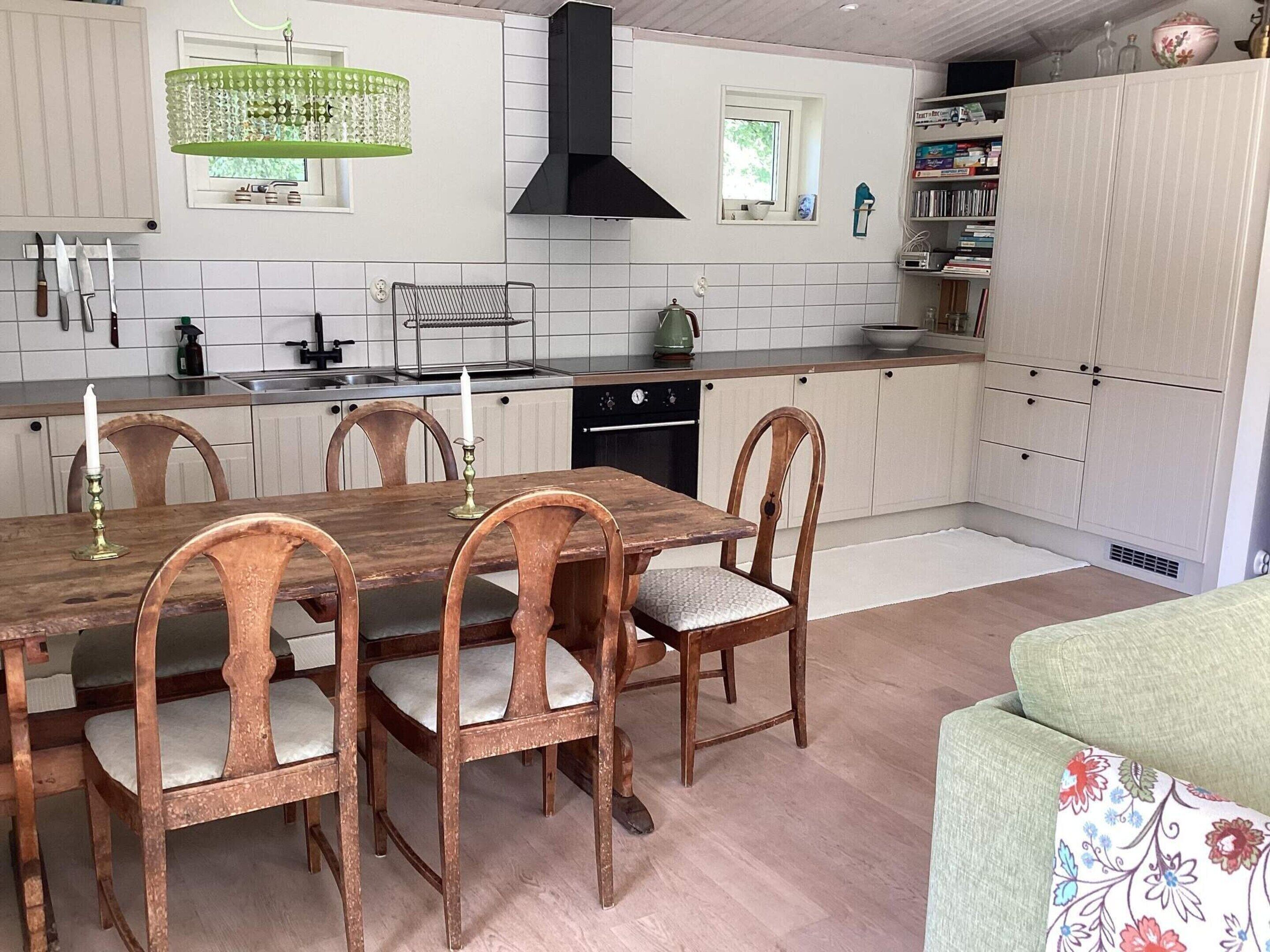Una cocina con una mesa y sillas de madera, una lámpara de araña verde, una campana extractora negra y paredes con azulejos blancos.
