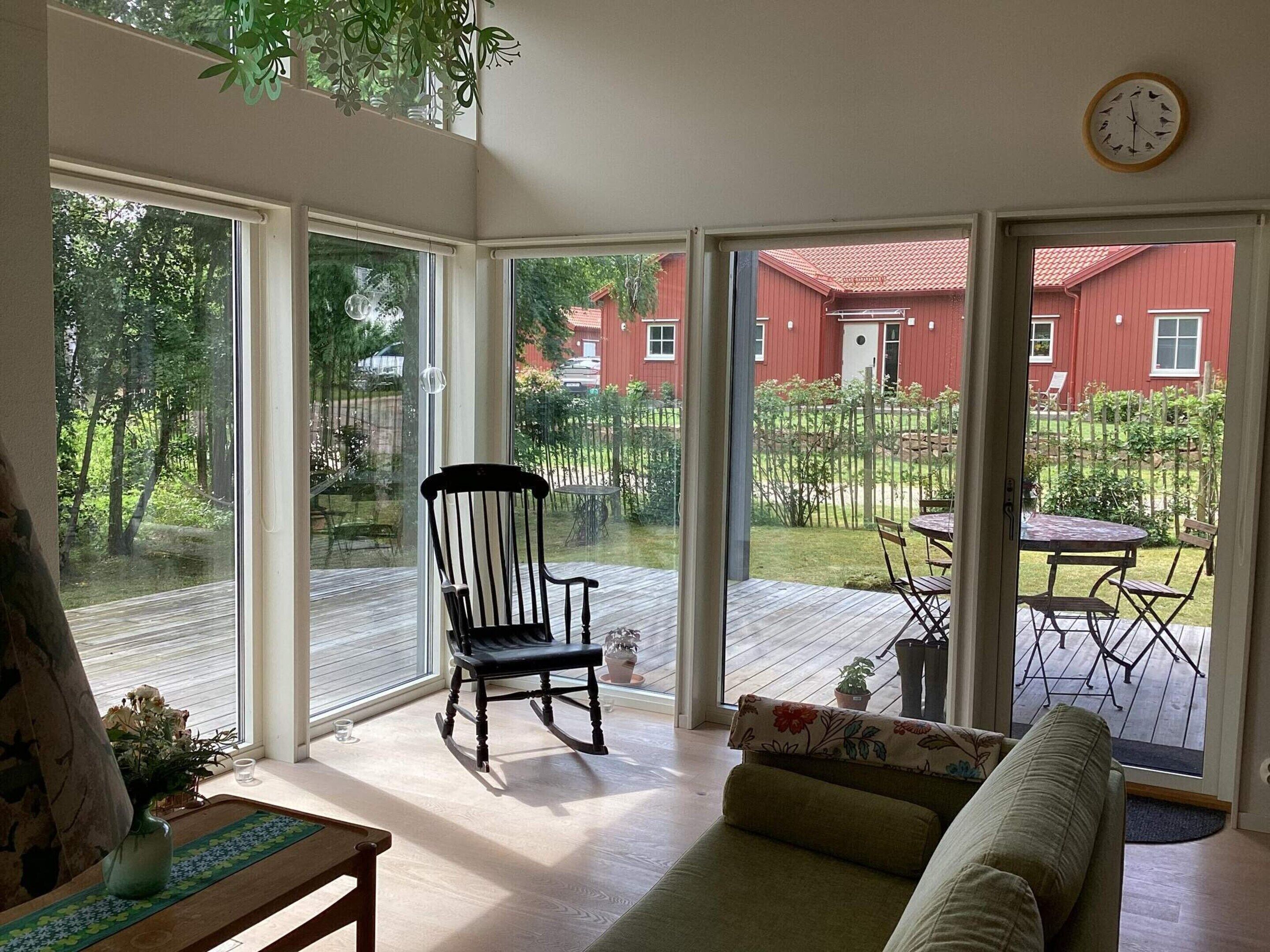 A room with a wooden floor, a rocking chair, a sofa, a table, and a view of a red building and greenery outside.