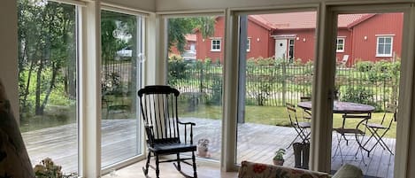 A room with a wooden floor, a rocking chair, a sofa, a table, and a view of a red building and greenery outside.