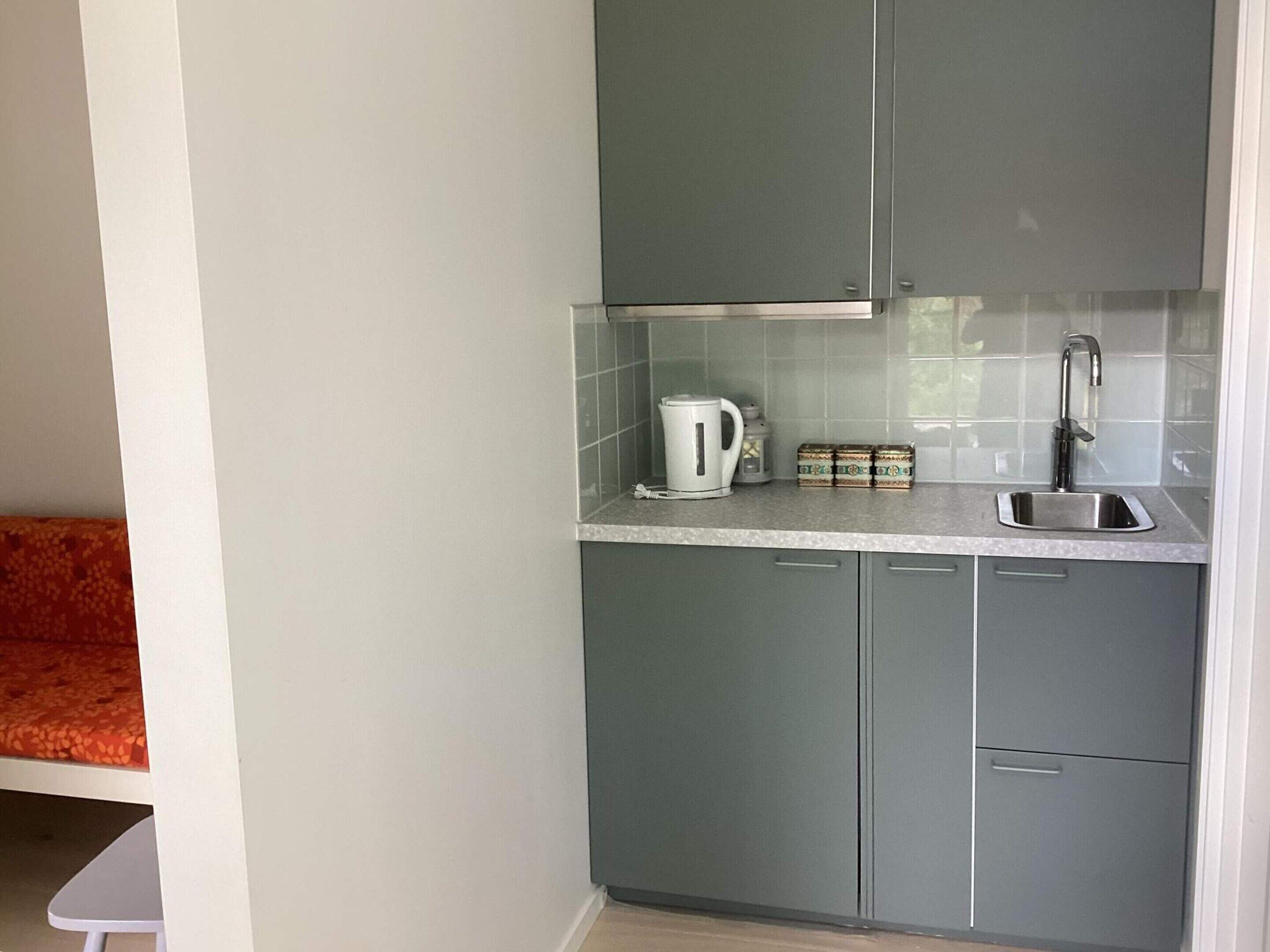 Una cocina compacta con gabinetes grises, una encimera blanca, una tetera, una caja pequeña y un lavabo.