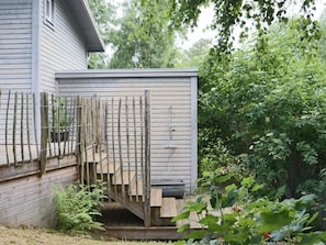 Eine Holzdielen-Terrasse mit einem Metallzaun, ein Haus mit grauer Fassade und üppiges Grün im Hintergrund.