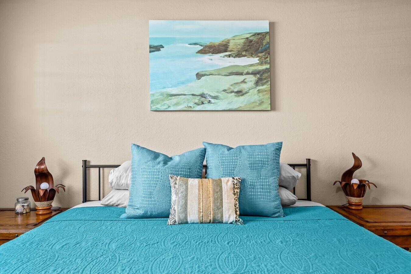 Una cama con una colcha color aguamarina, dos almohadas y un cuadro de un paisaje costero encima.