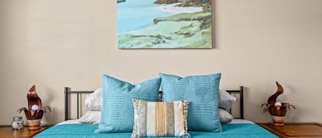 A bed with a teal bedspread, two pillows, and a painting of a coastal landscape above it.