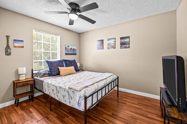 A bedroom with a bed, a ceiling fan, a window with blinds, a bedside table, a TV stand, and wall art.